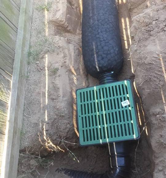 Narrow trench beside a wooden retaining board with a green grated catch basin, a black drain line wrapped in a filter sock above it and drain pipe below.