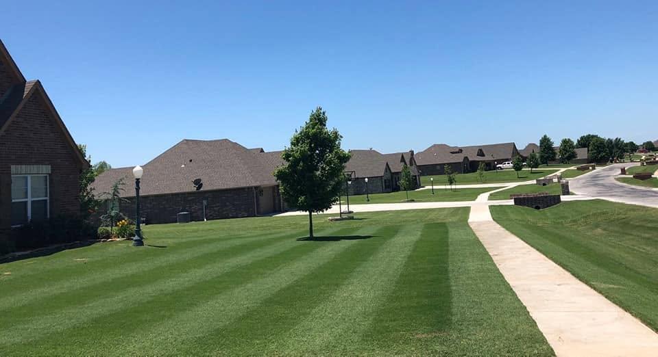 Mown lawn with stripes running toward a brick house with a lamp post, a sidewalk curving along the edge, under a bright blue sky.